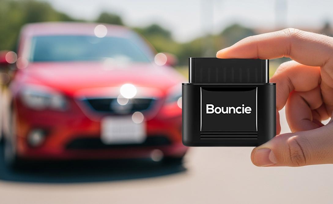 Hand holding a Bouncie tracking device in front of an out-of-focus red car